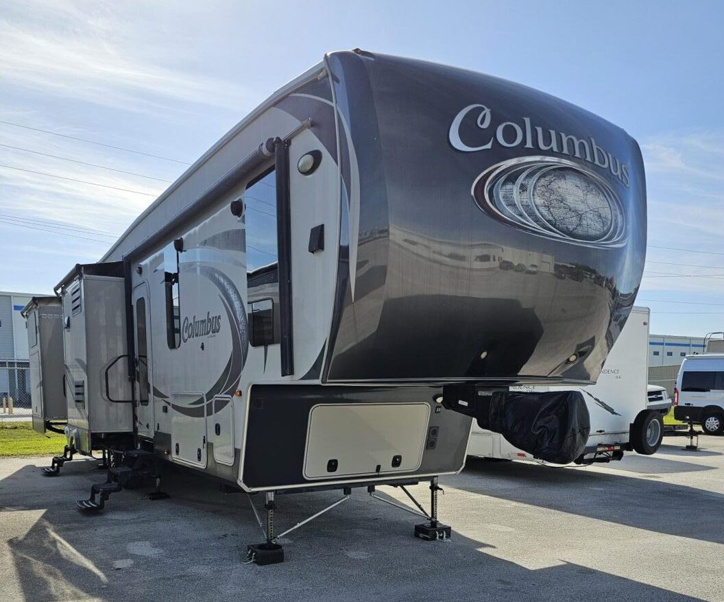 A large columbus fifth-wheel rv parked on a concrete lot with extended slide-outs and a prominent logo on the front cap.